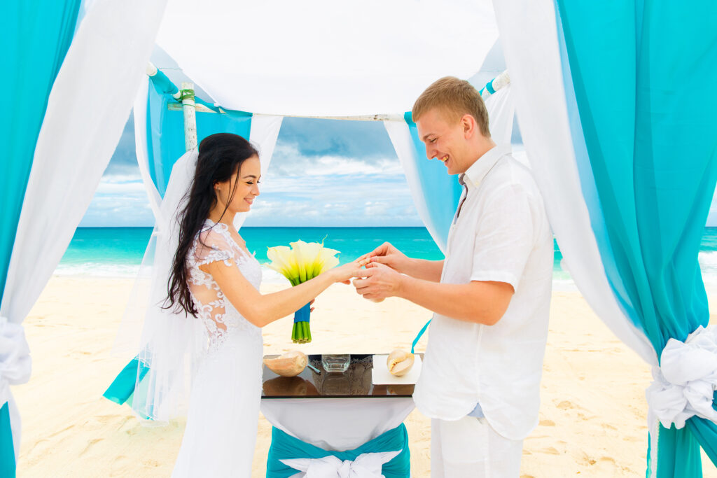 Groom giving an engagement ring to his bride under the arch decorated with flowers on the sandy beach. Wedding ceremony on a tropical beach in blue. Wedding and honeymoon concept.